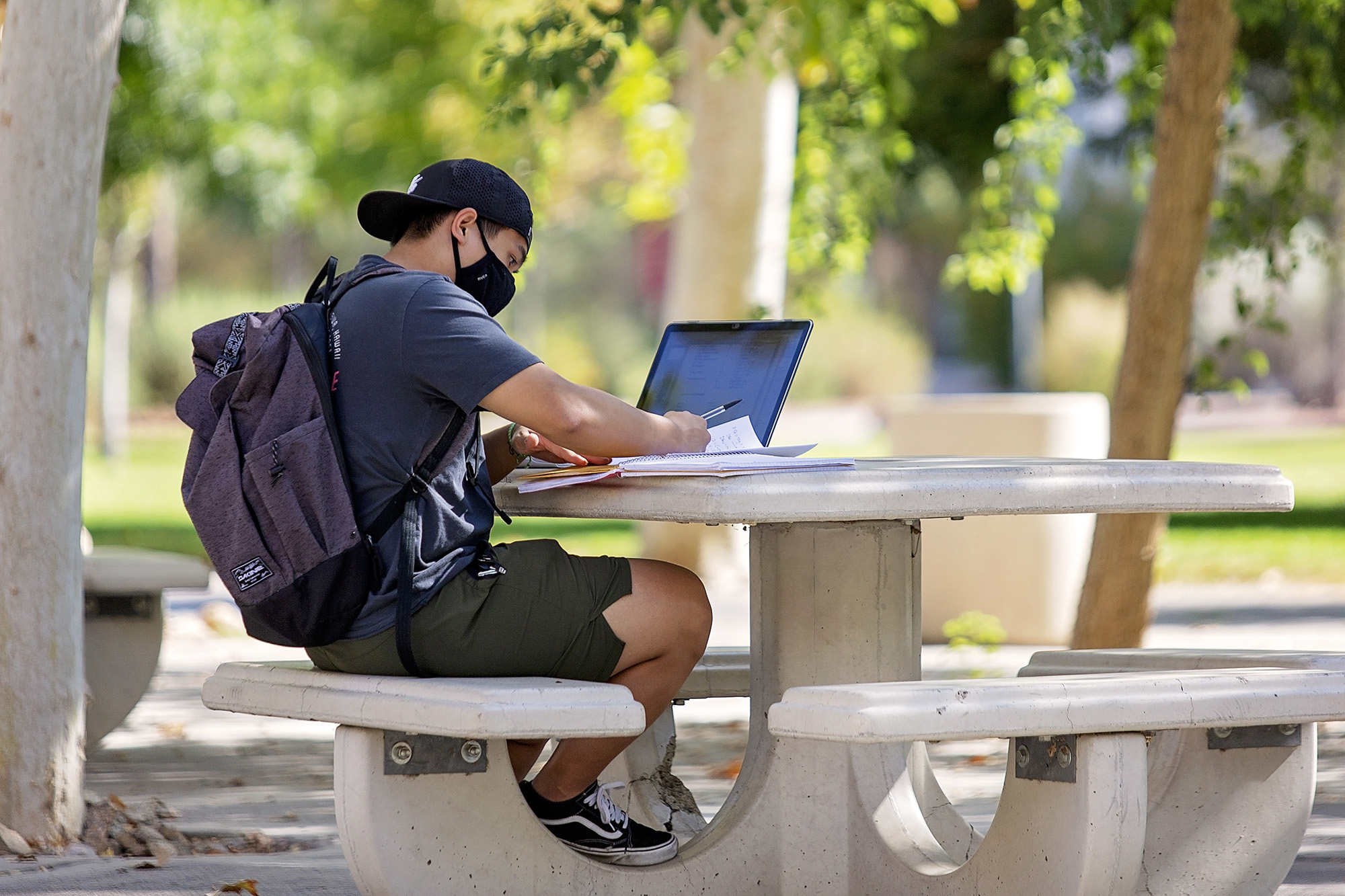 A masked student sits at an outdoor table doing schoolwork.