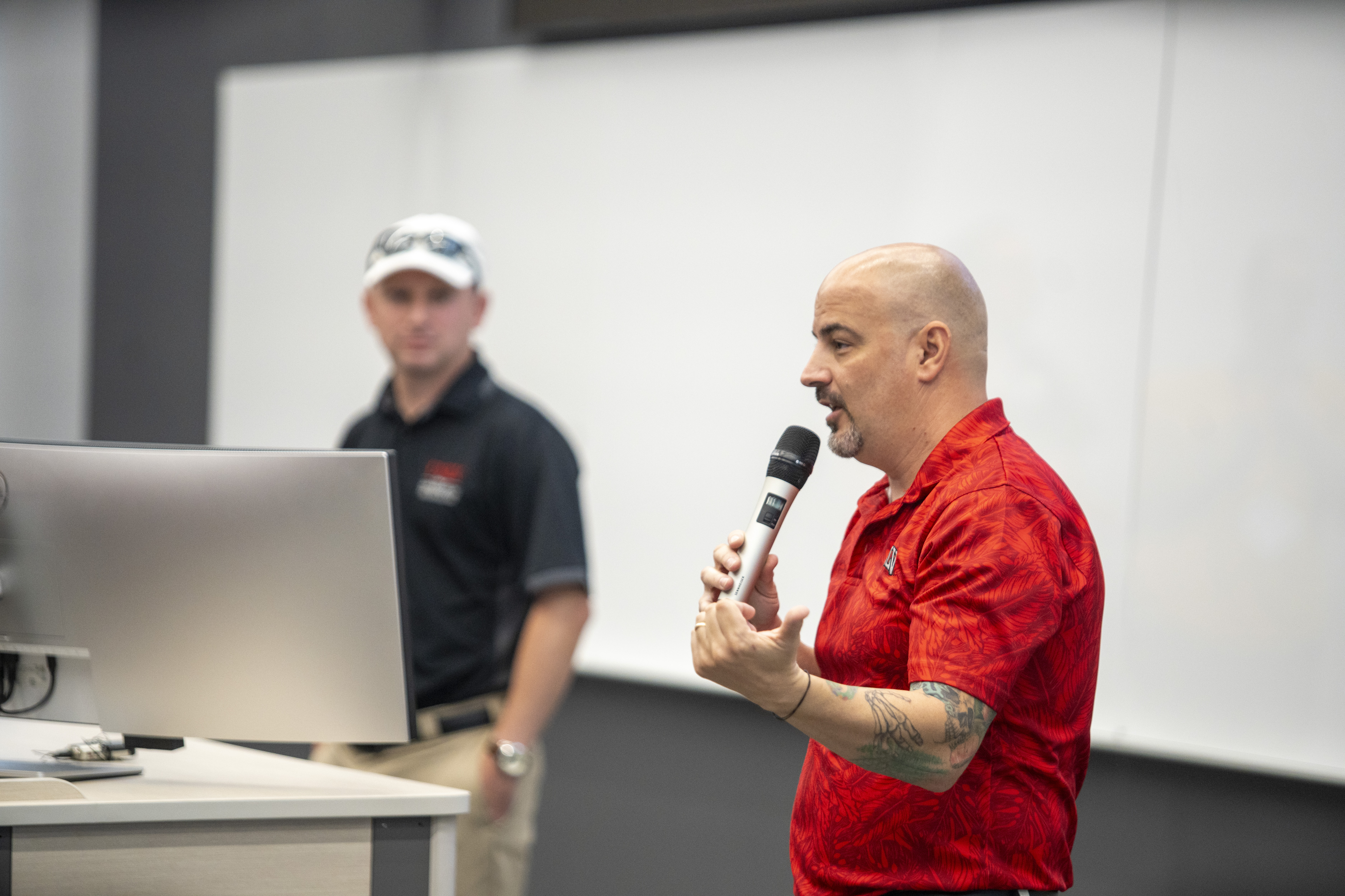Person holding a microphone and talking with another person in the background