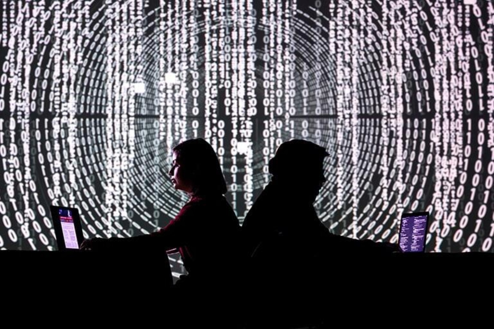 Two people on laptop with code on wall behind them.