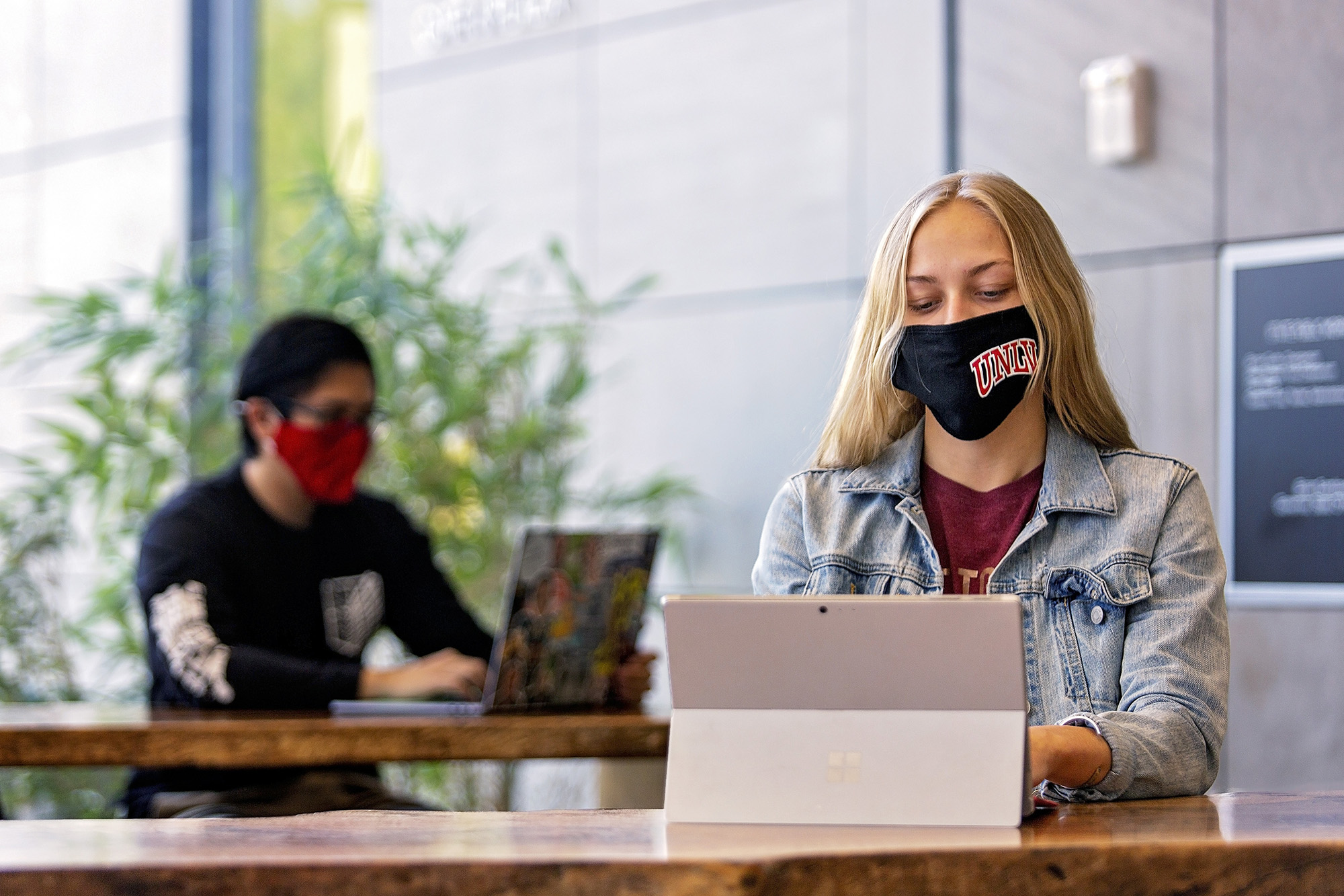 A masked student sits at a wood table studying on a tablet.