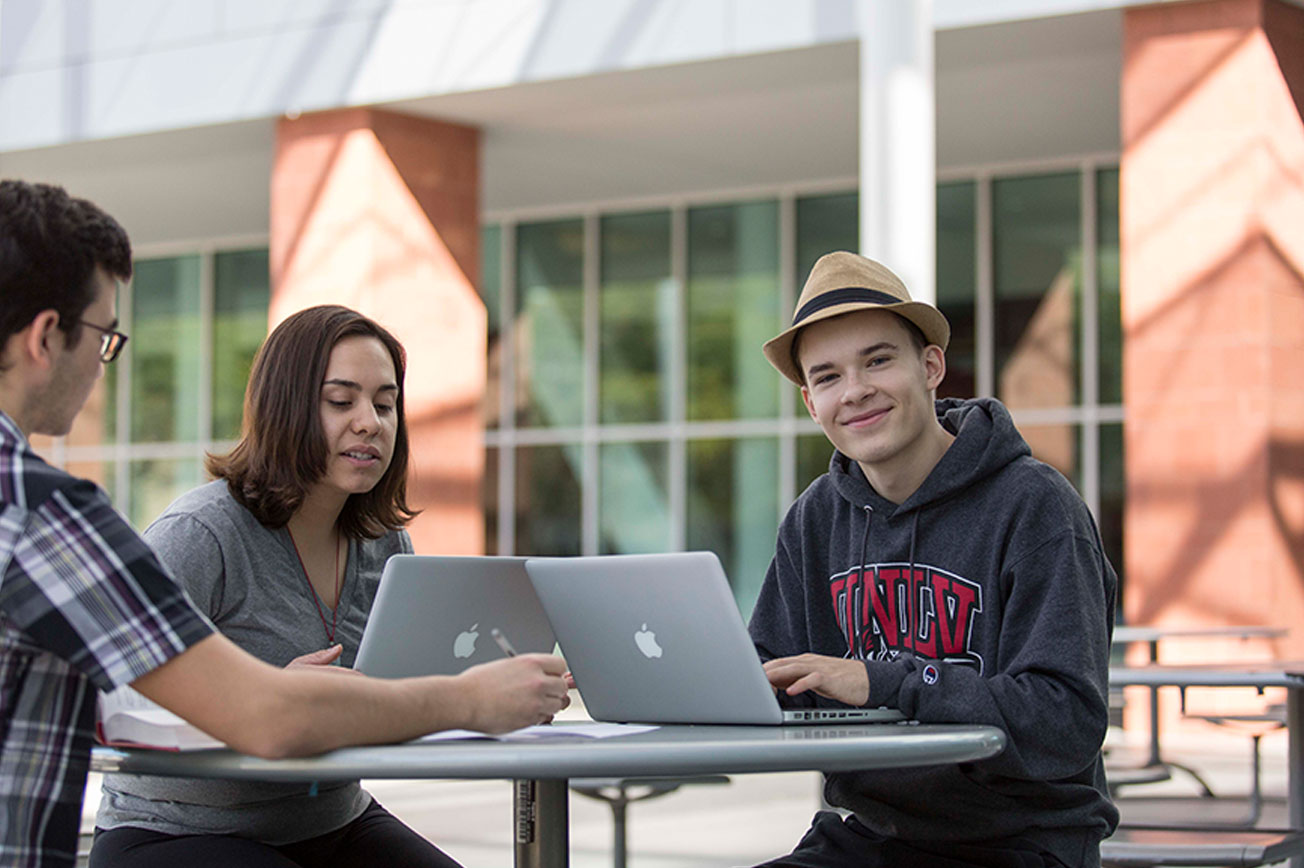 A male student smiles at the camera while he studies at an outdoor table with two other students.