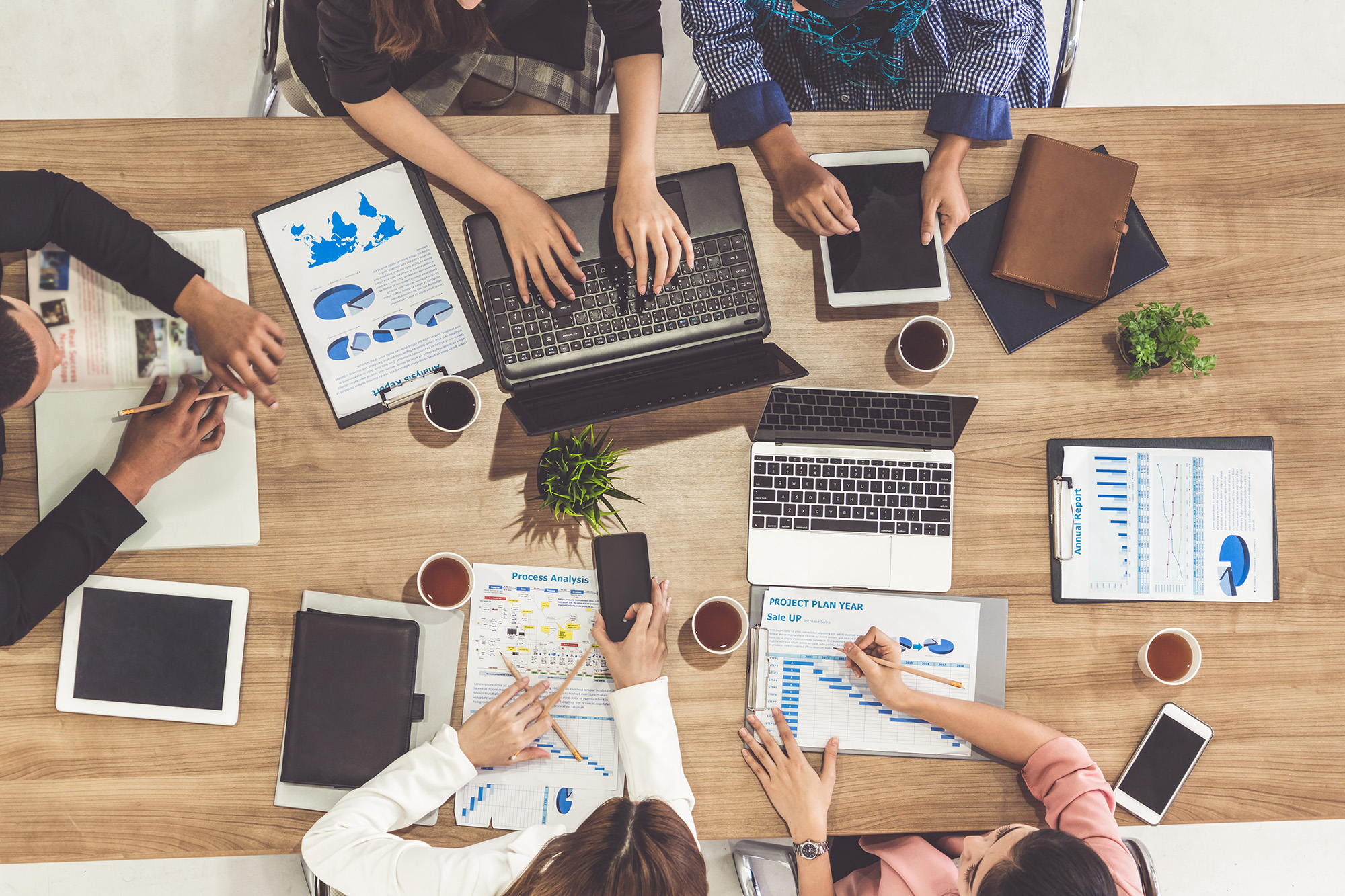 Top-down view of a diverse team working together at a table with laptops, tablets, reports, and coffee, discussing data and project plans.