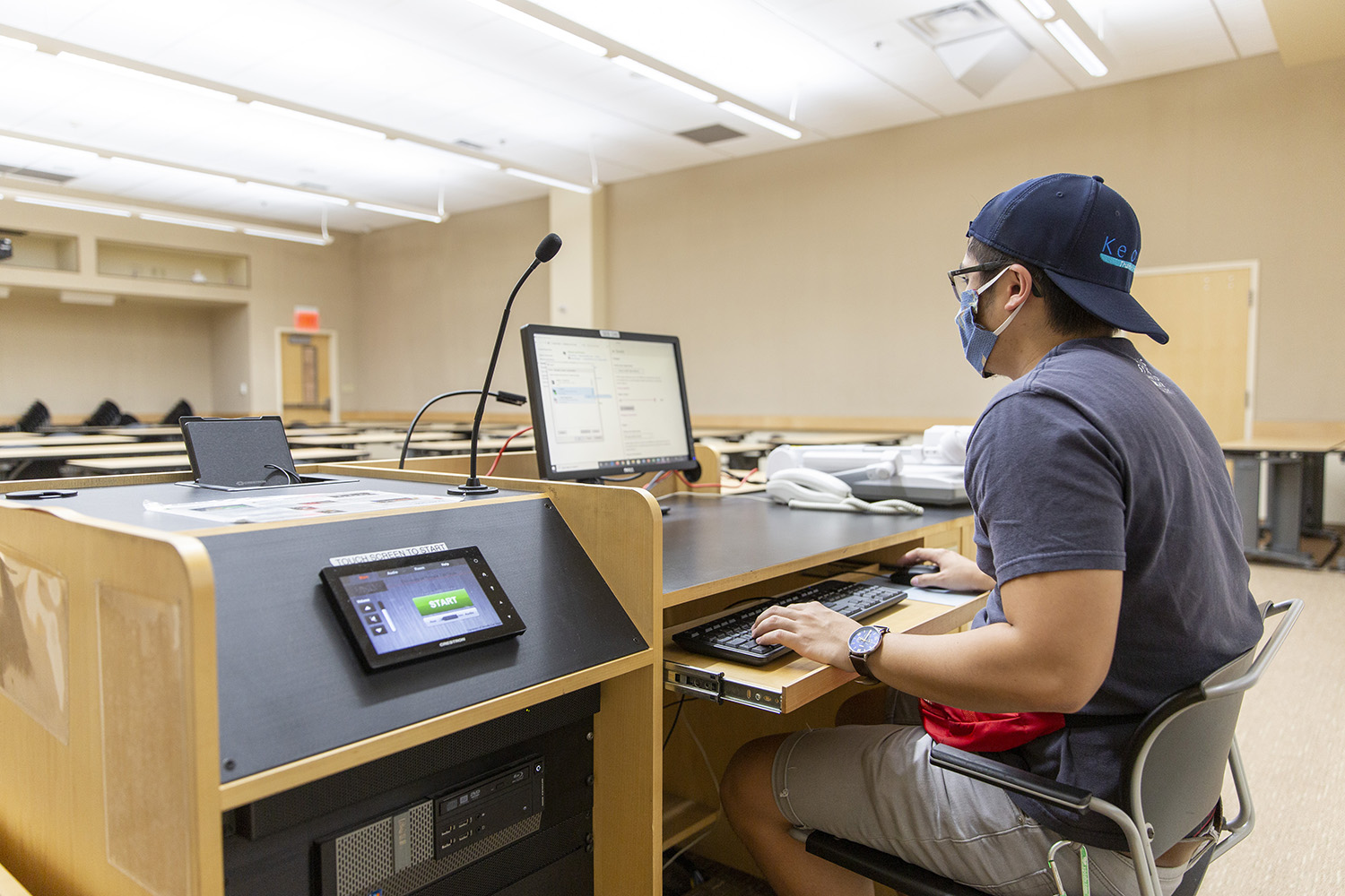 Staff member sitting behind lectern using desktop computer in a classroom.