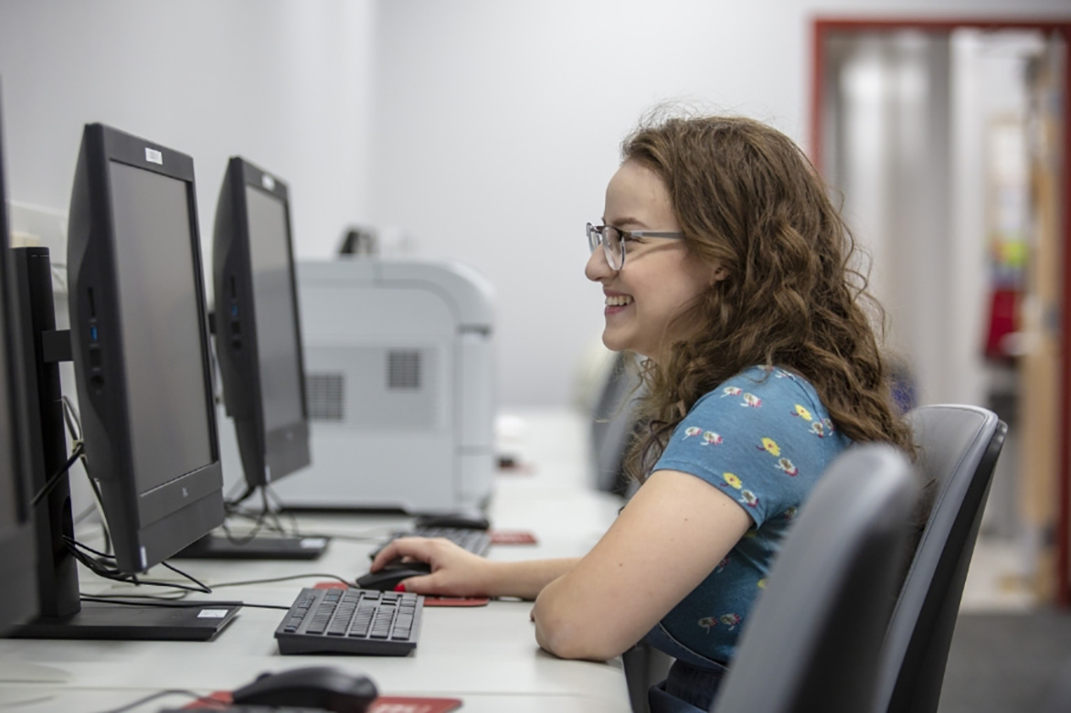 A smiling female student sits in front of a computer lab workstation.