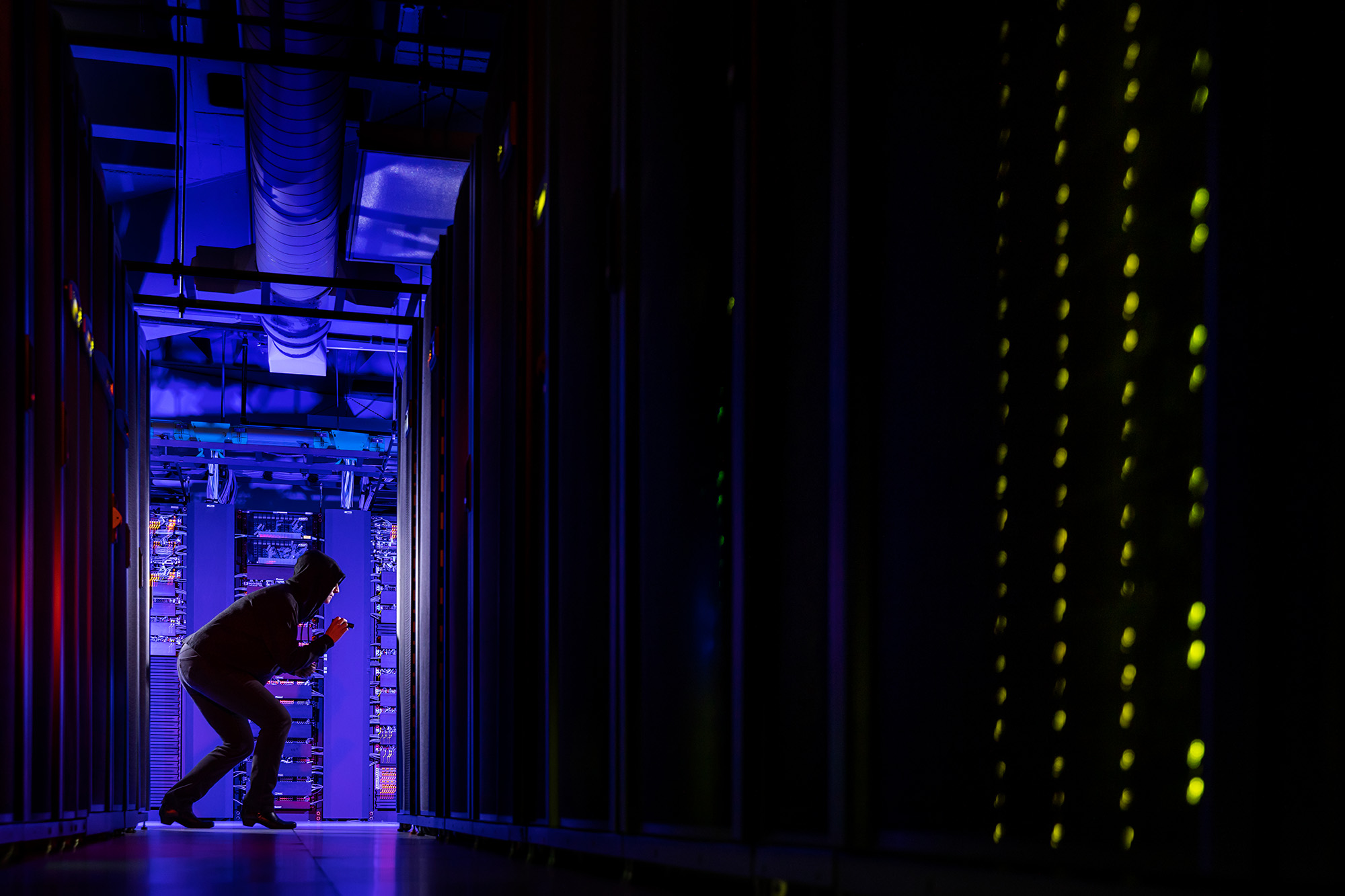 A black-hooded figure with a small flashlight appears to be snooping in a data center.