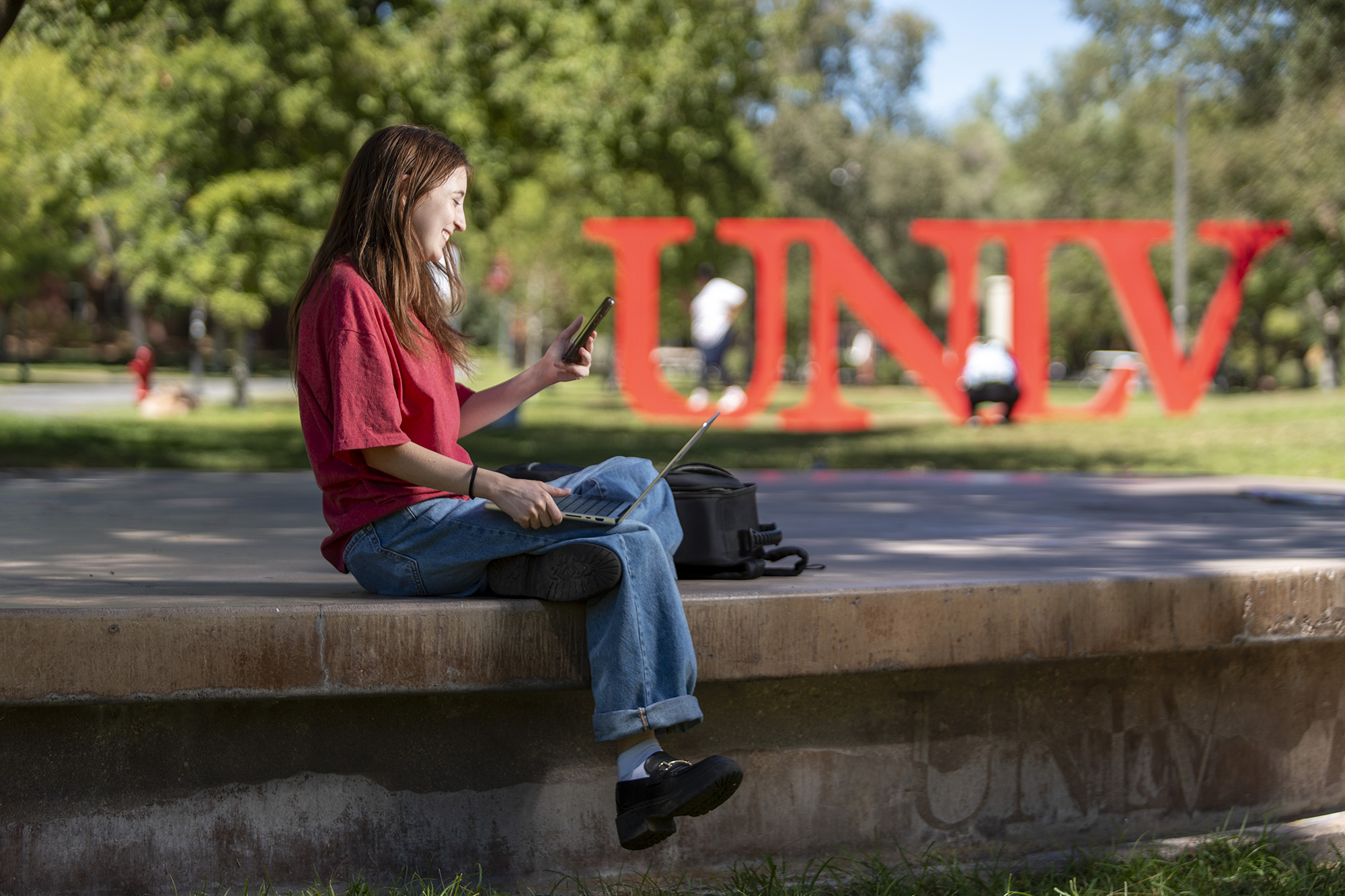 A student posing with RebelCard student ID and laptop in front of red U-N-L-V marquee letters.