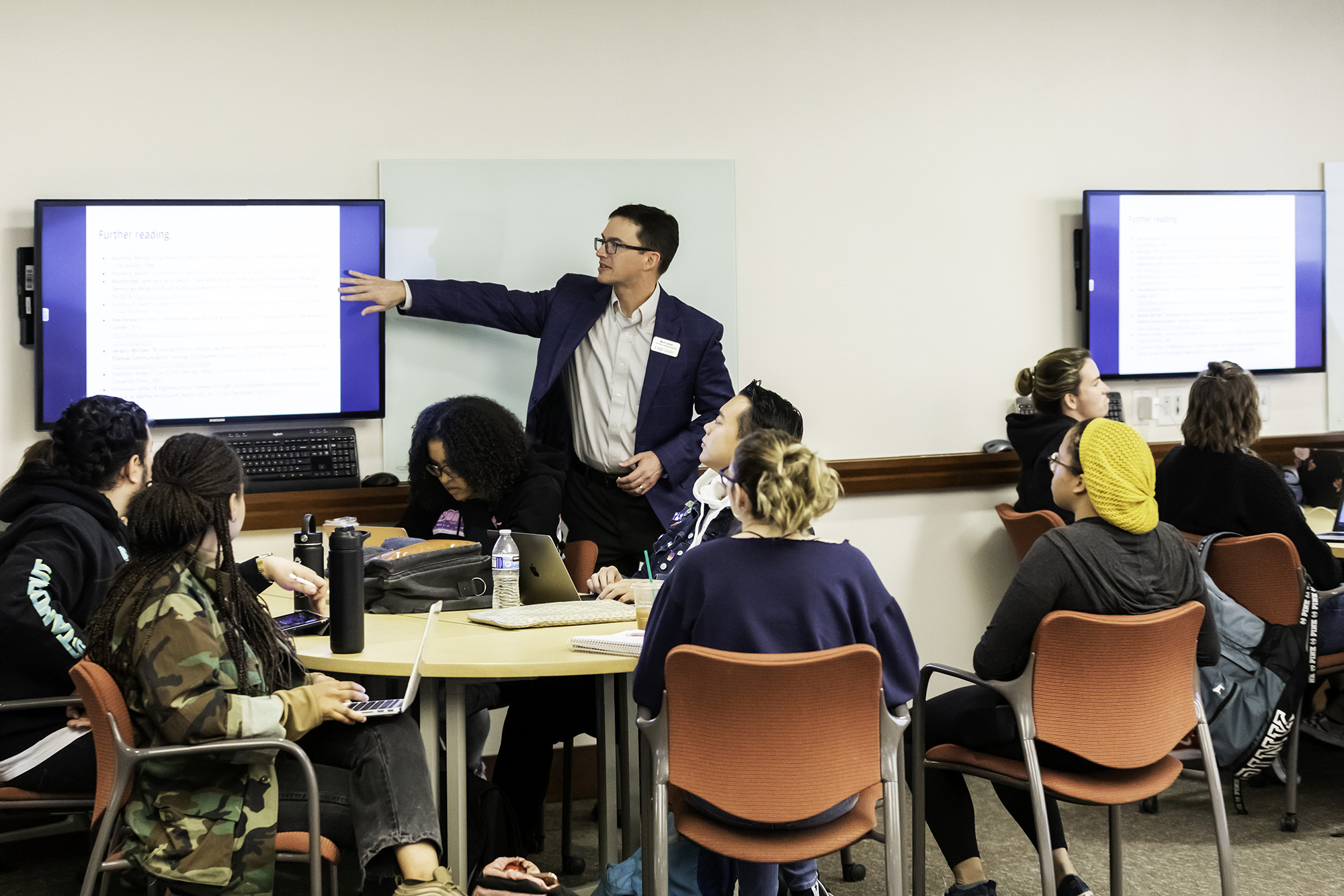 An instructor points to a wall-mounted monitor as students look on.