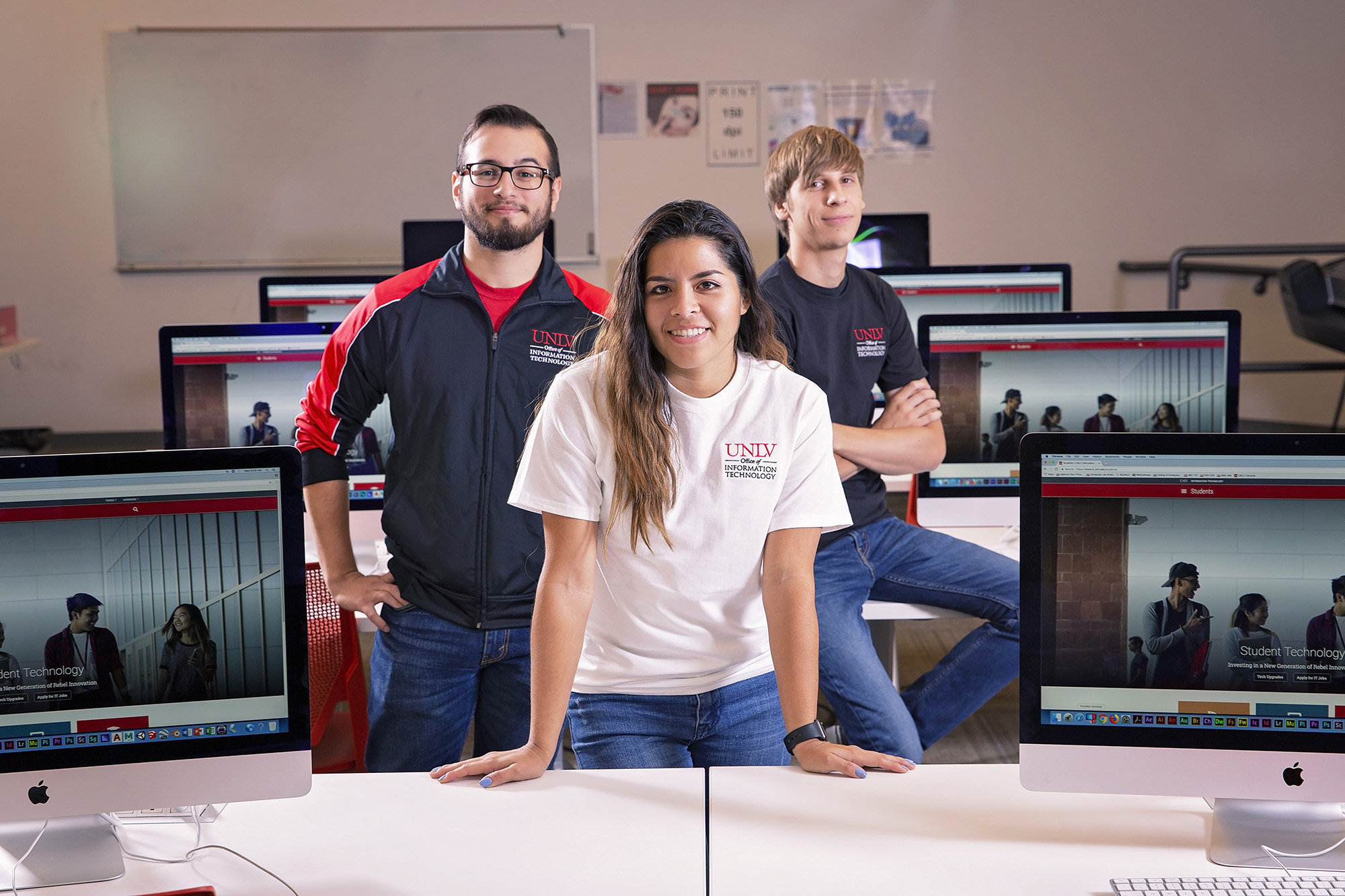 Three students pose in between a row of MacBooks.