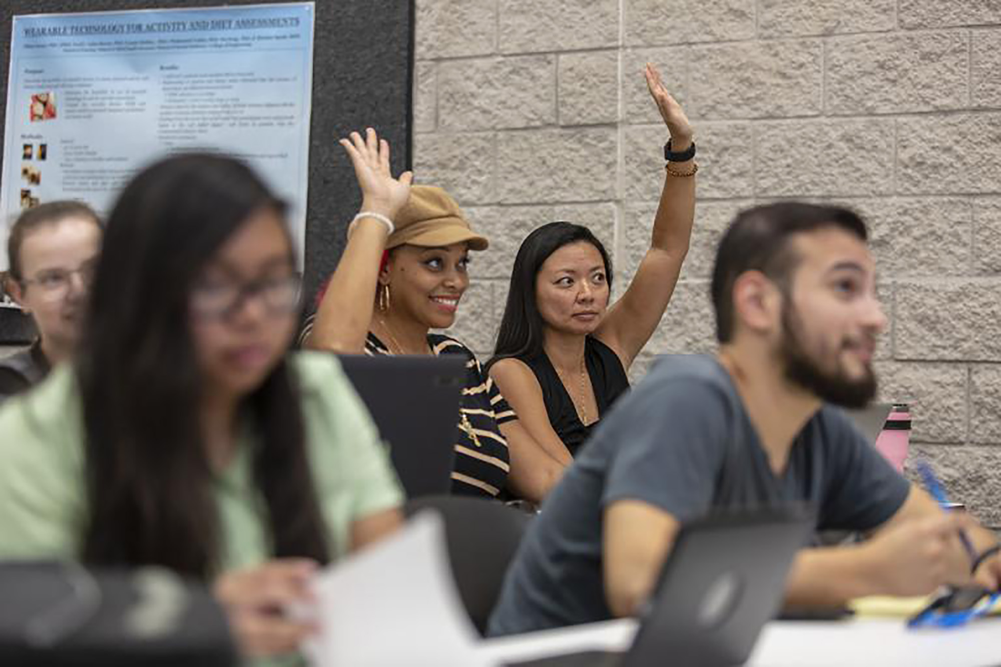 Two female staff members in a classroom raise their hands.