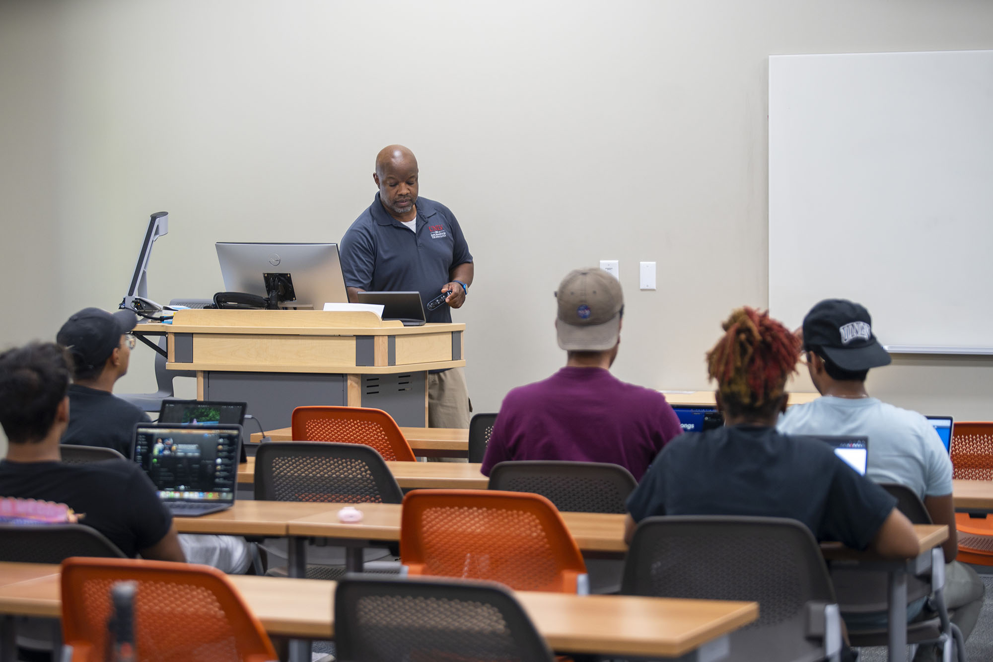 Instructor standing behind lectern in front of classroom with students using laptop computers.