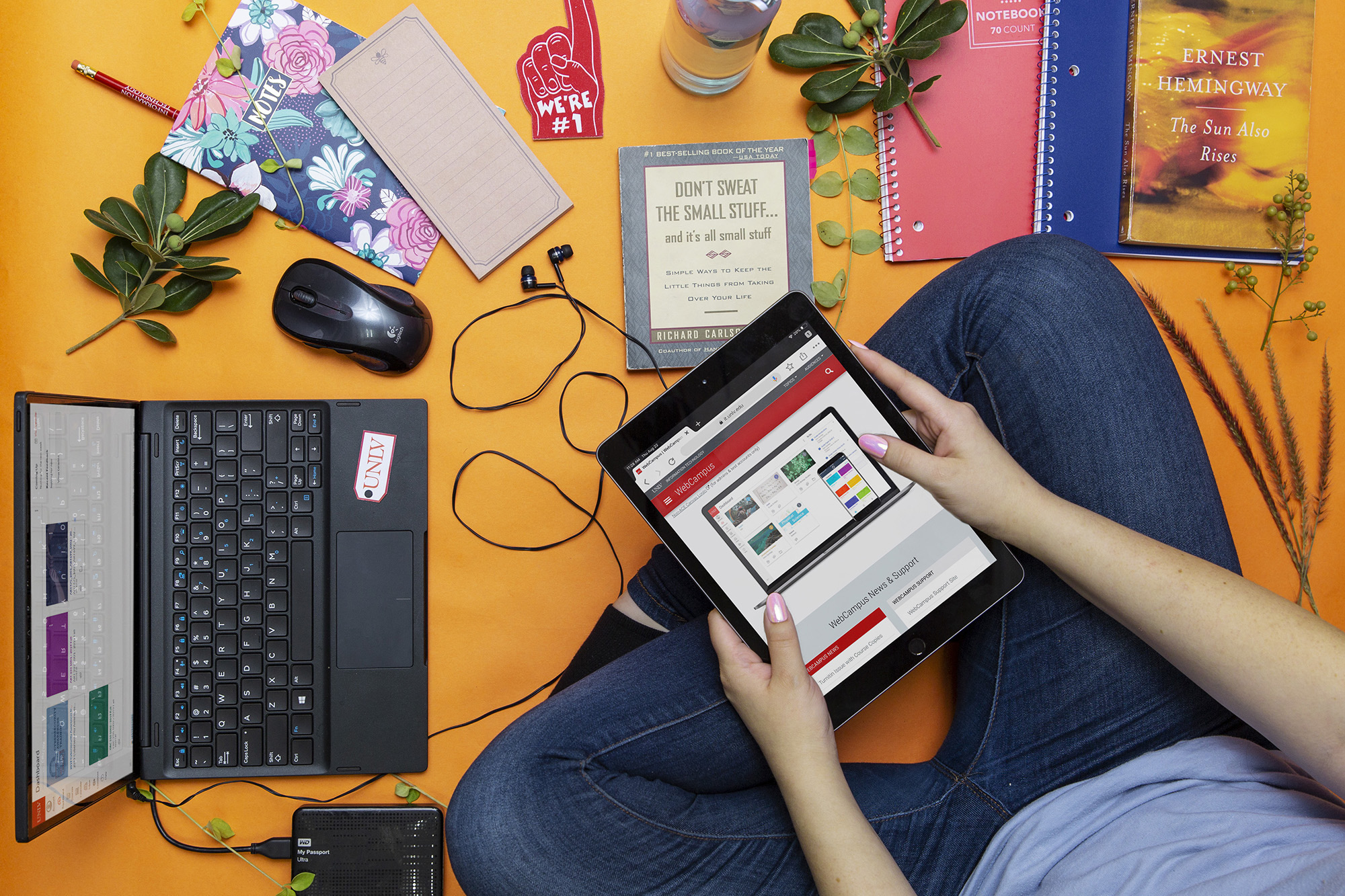 A female student, surrounded by a laptop and various school supplies, holds a tablet displaying the WebCampus web page.