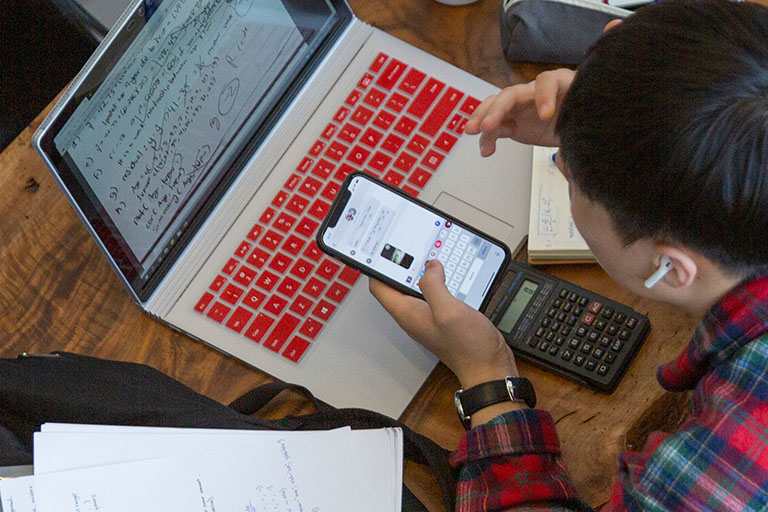 A male student types on his mobile device with an open laptop in front of him.