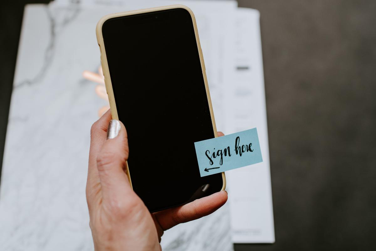 Hand holding a smartphone with a sticky note that says "Sign Here" afixed.