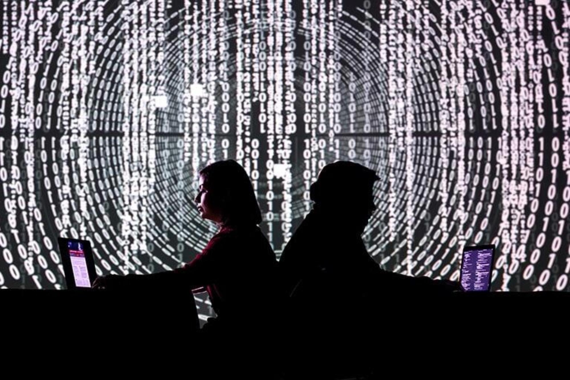 Two people on laptop with code on wall behind them.