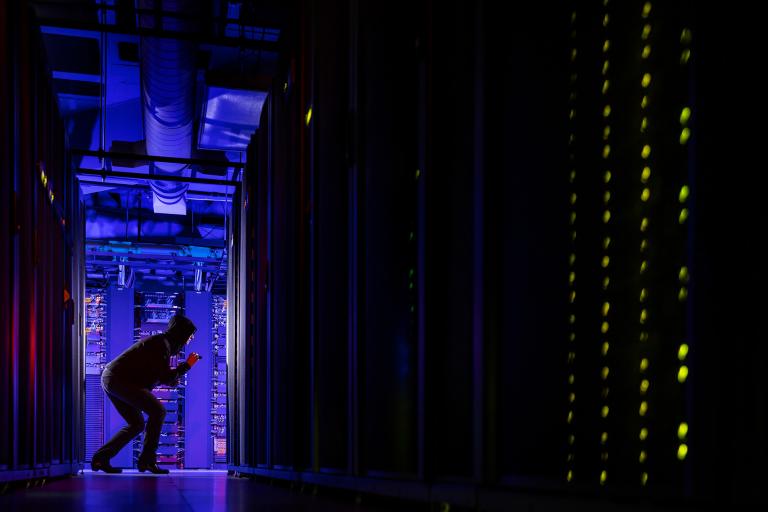A black-hooded figure with a small flashlight appears to be snooping in a data center.