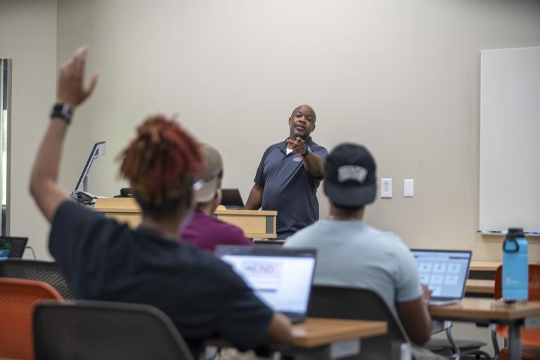 College instructor engaging with students in a modern classroom; one student has their hand raised while others work on laptops.