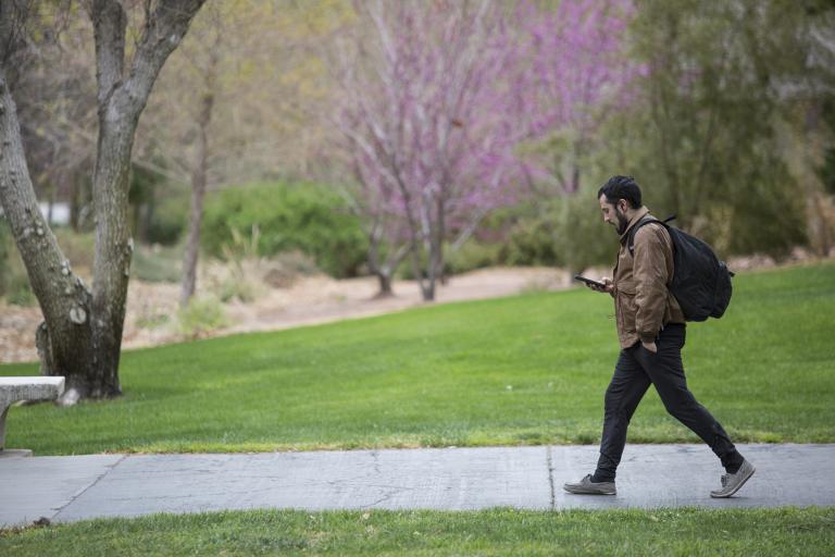 A male student looks at his smart phone while walking a university pathway.