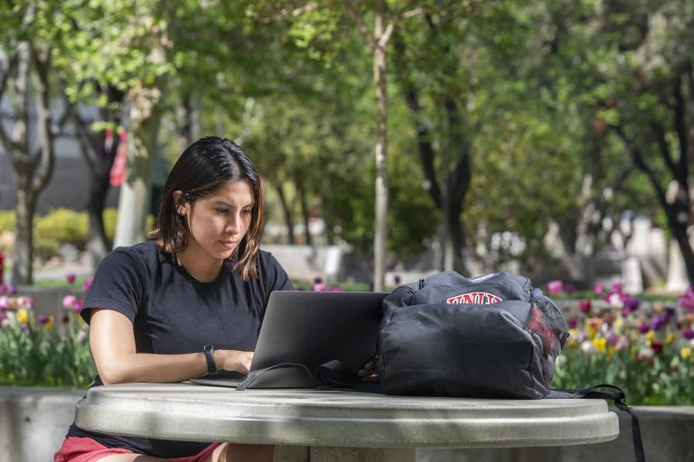 A student with a laptop sitting outdoors with a rose garden in the background.