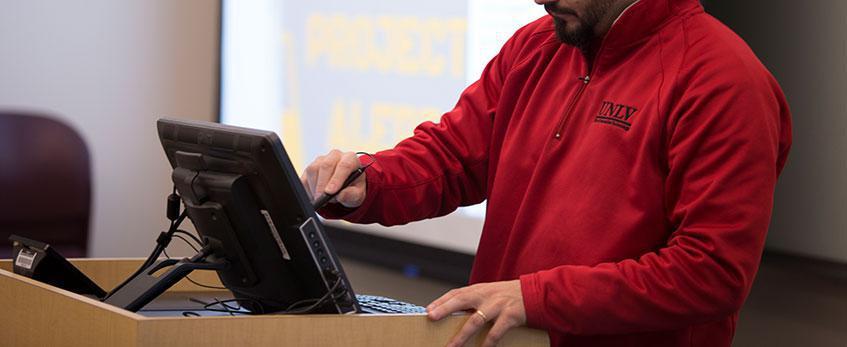 A male staff member standing at a lectern sets up classroom technology.