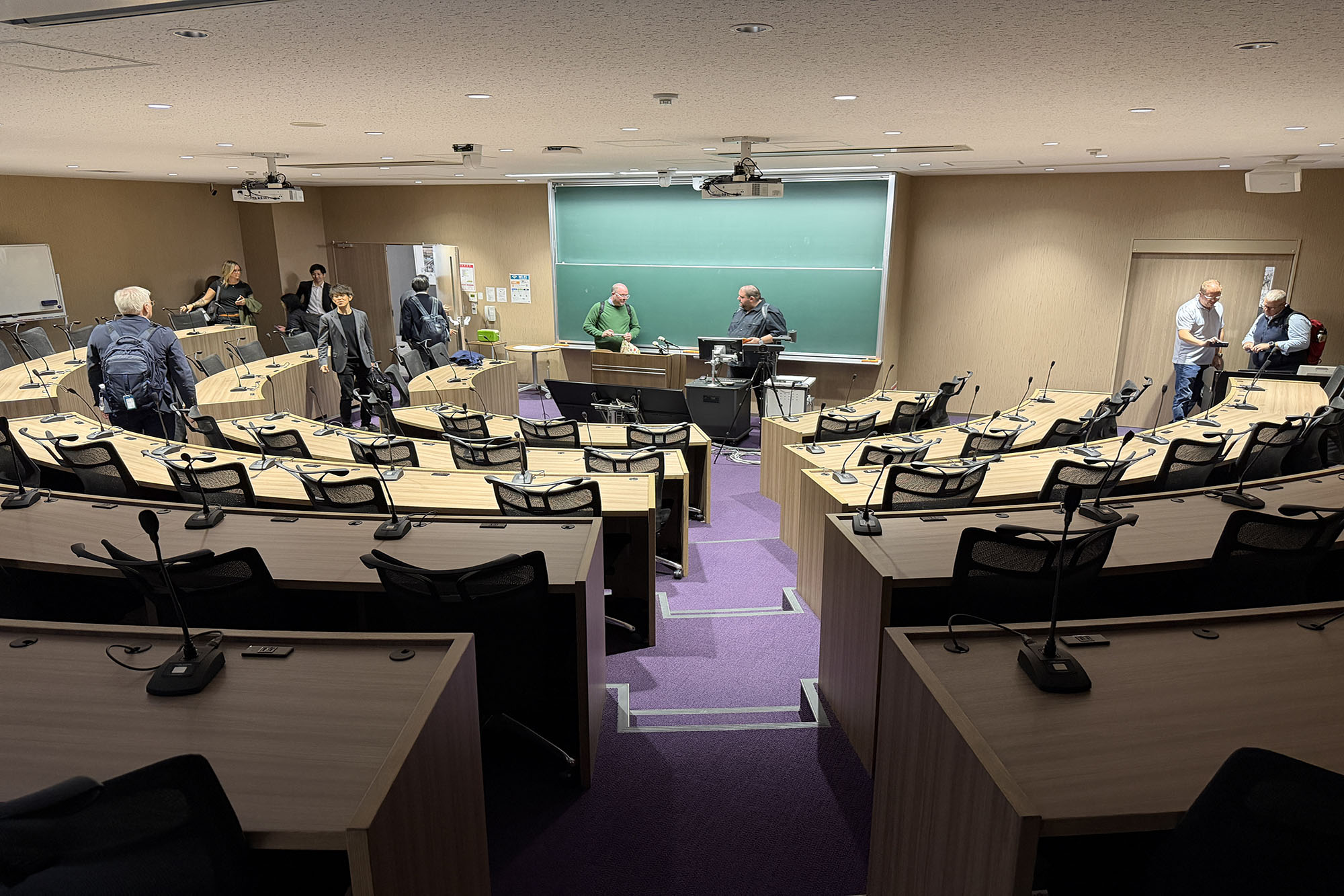 Tiered lecture hall classroom with curved rows of desks, microphones, and computer stations facing a large chalkboard and presentation area, while several people stand and talk around the room.